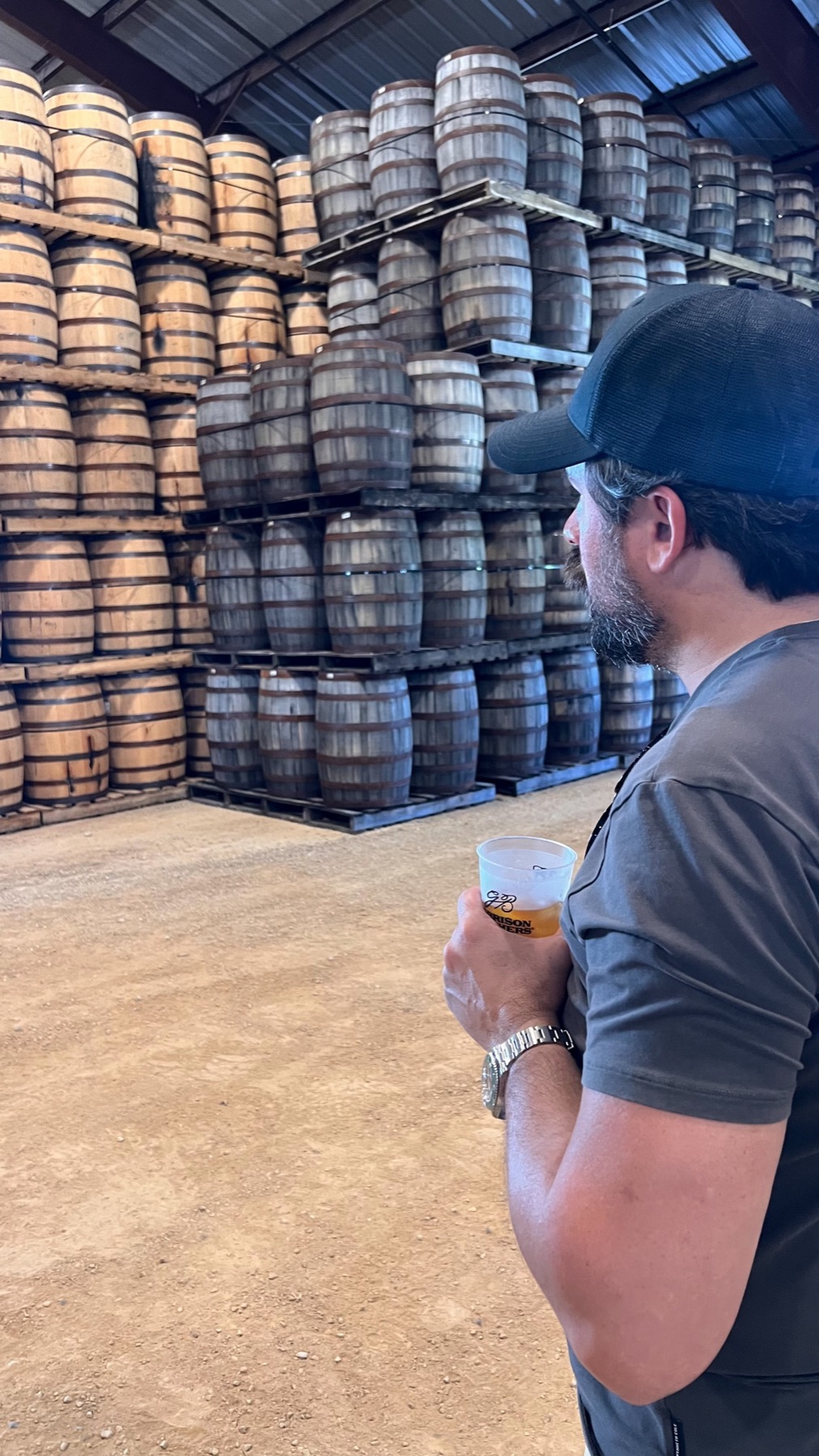 Matt in profile holding a tasting cup in front of a wall of stacked barrels in the rickhouse