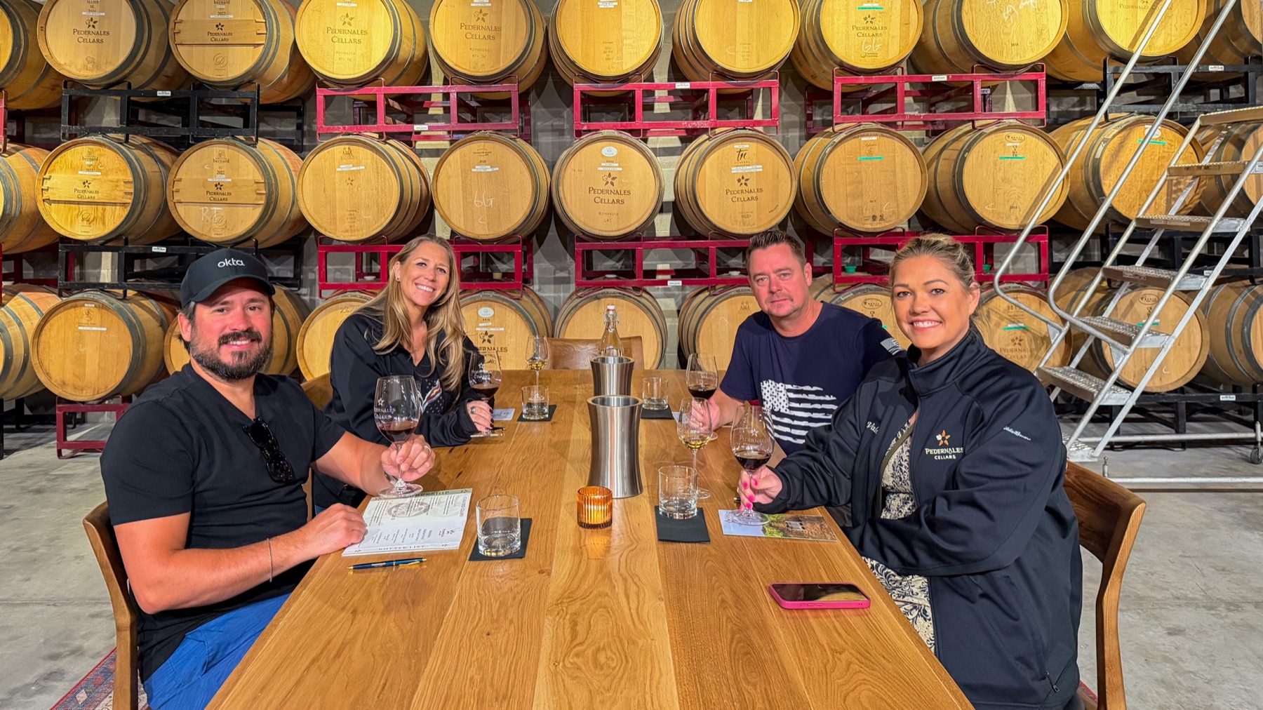 All four seated at a long wooden tasting table in front of a wall of barrels, wearing lent jackets