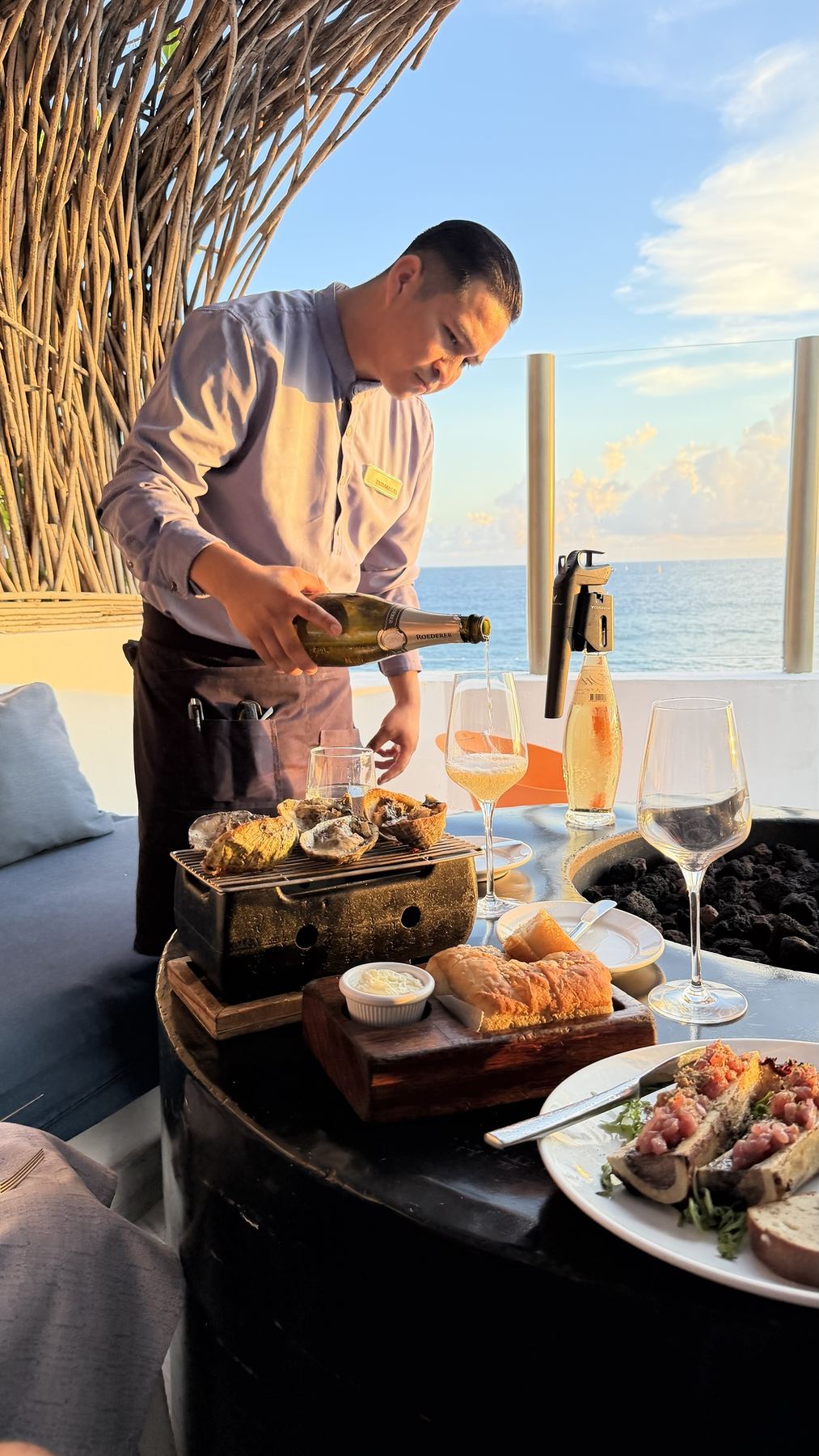 A waiter pouring Roederer champagne beside grilled oysters and bone marrow, ocean behind