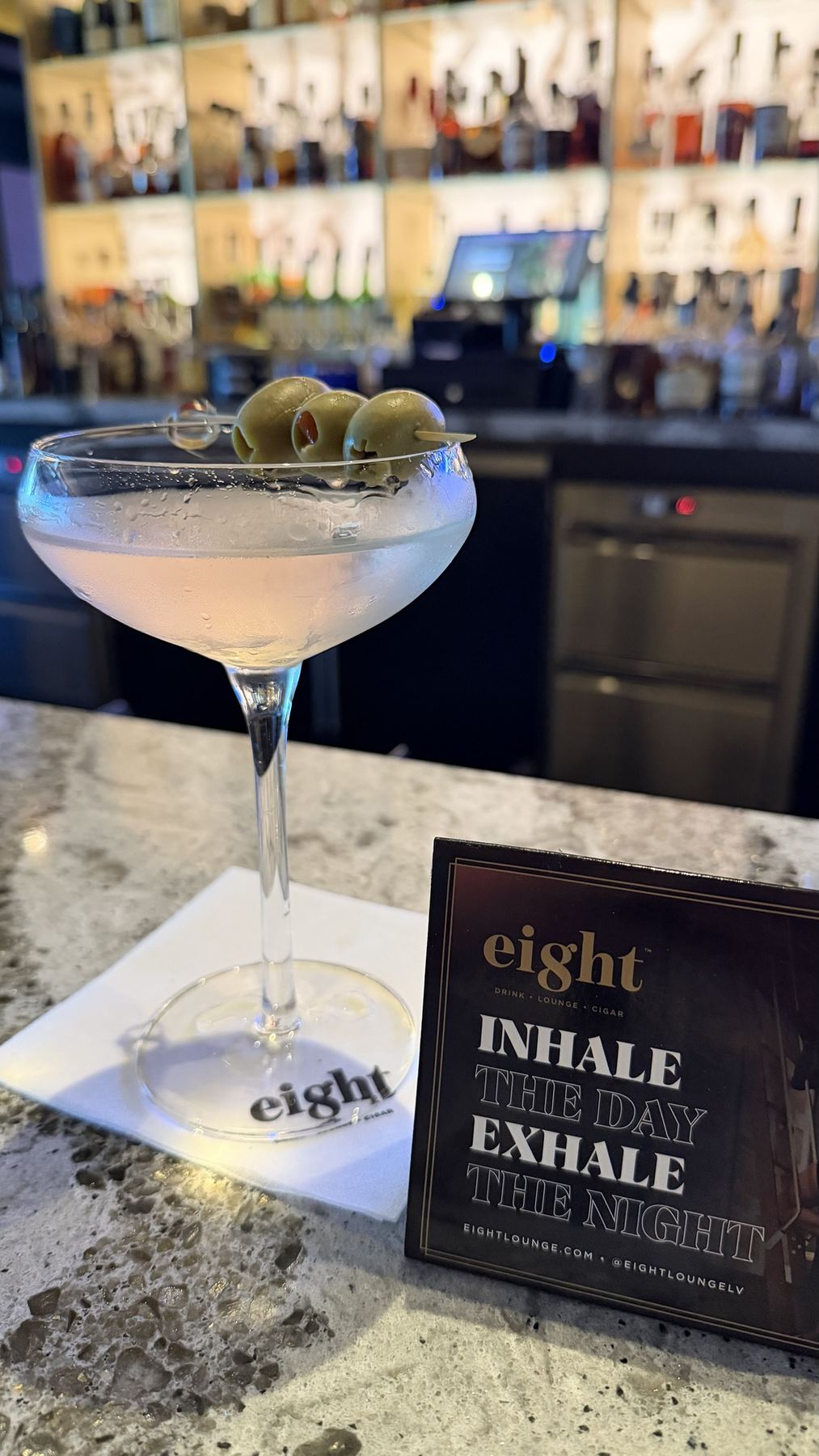 A frosted martini with three olives beside a table card reading inhale the day, exhale the night